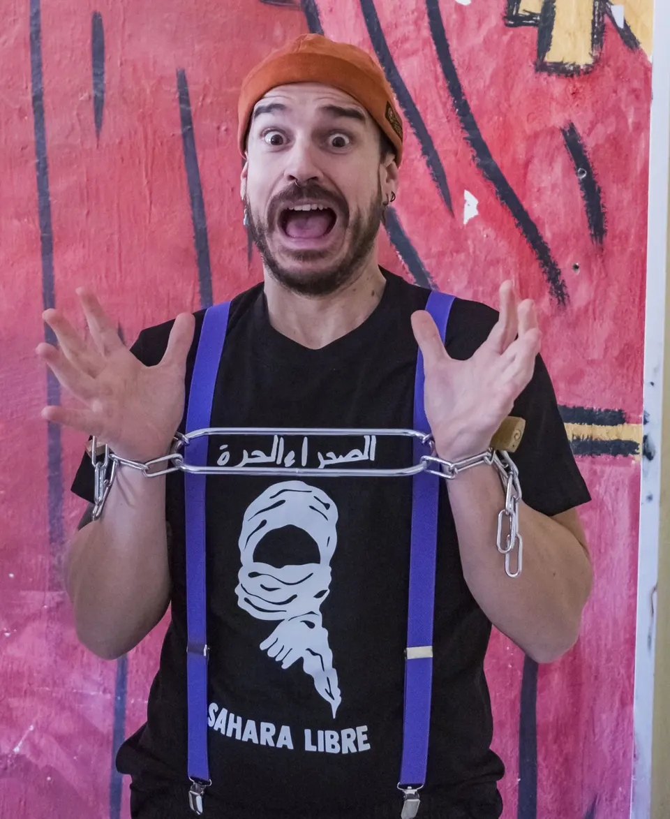 Un mago con camiseta de «Sahara Libre» y cadenas en las muñecas pone cara de sorpresa ante un mural rosa