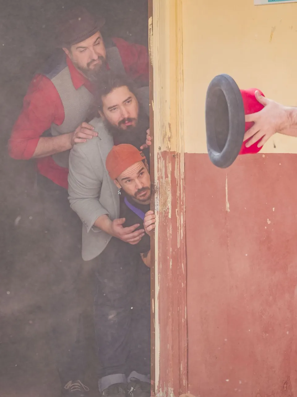 Tres magos asoman uno encima de otro por el marco de una puerta mientras una mano lanza un sombrero